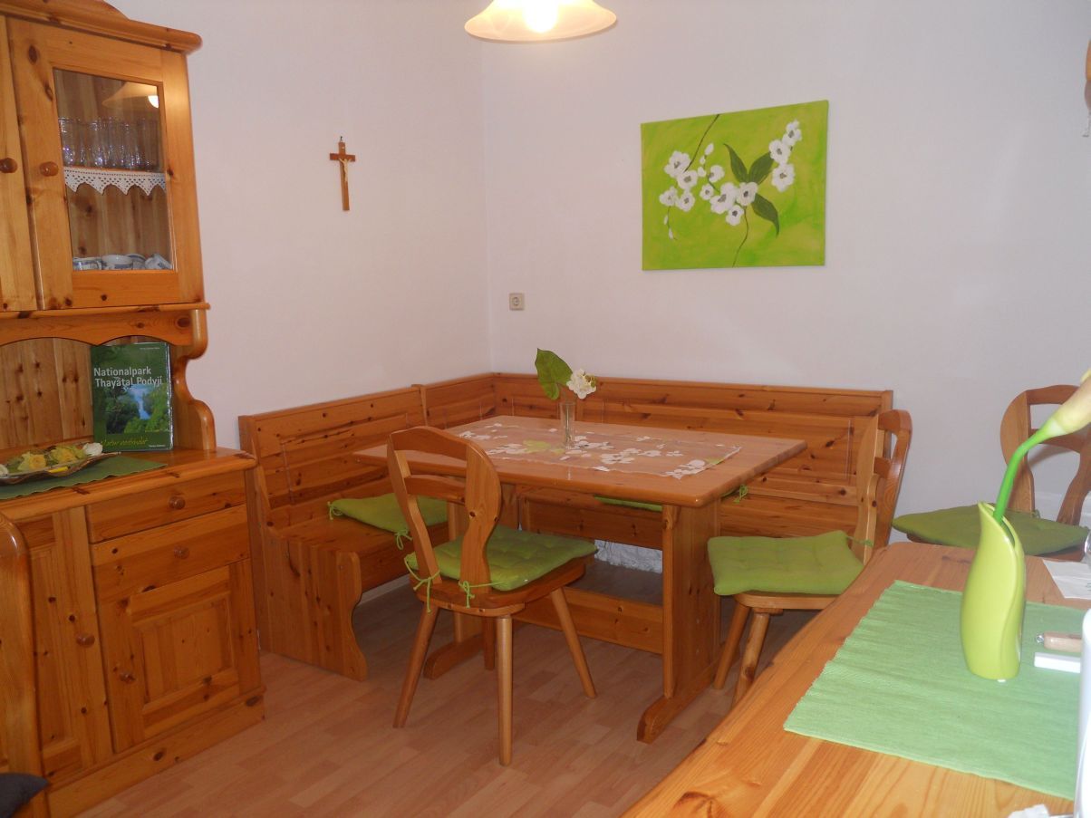 Gemütlicher Aufenthaltsraum mit Holzmöbeln, Eckbank, Tisch und Dekoration in Grün. An der Wand ein Kreuz und ein Bild mit Blumenmotiv.