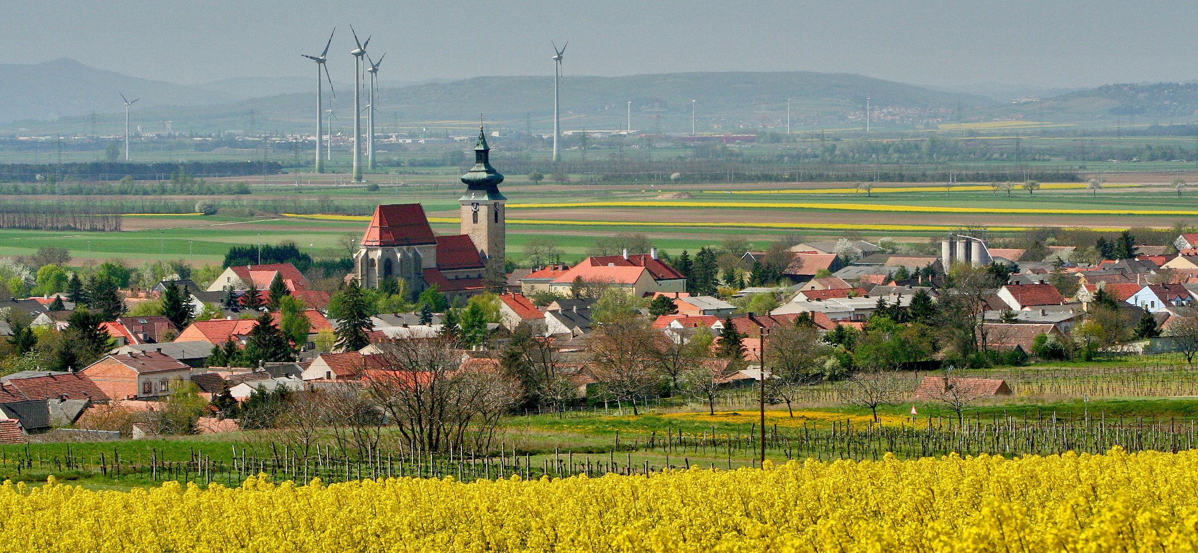 Landschaft mit Dorf, Kirche, Windrädern und Rapsfeld.