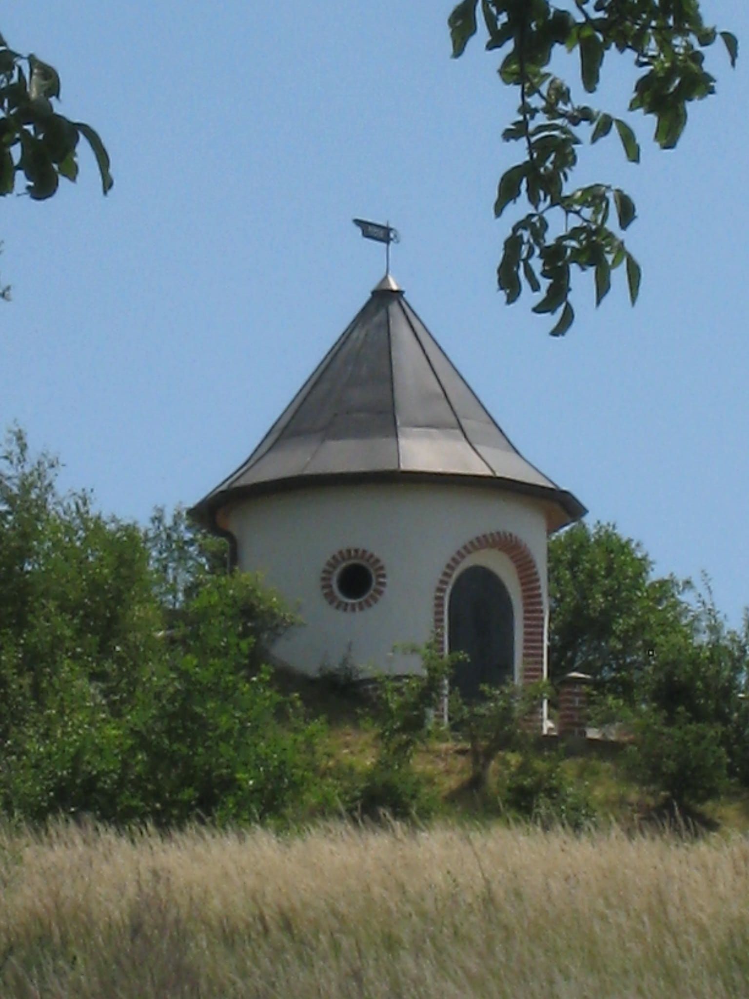 A round water tower with a pointed roof and weather vane, surrounded by trees and meadows.