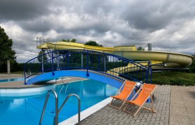 Zwei orange Liegest&uuml;hle stehen neben einem Schwimmbecken mit einer blauen Br&uuml;cke und einer gelben Wasserrutsche im Hintergrund.