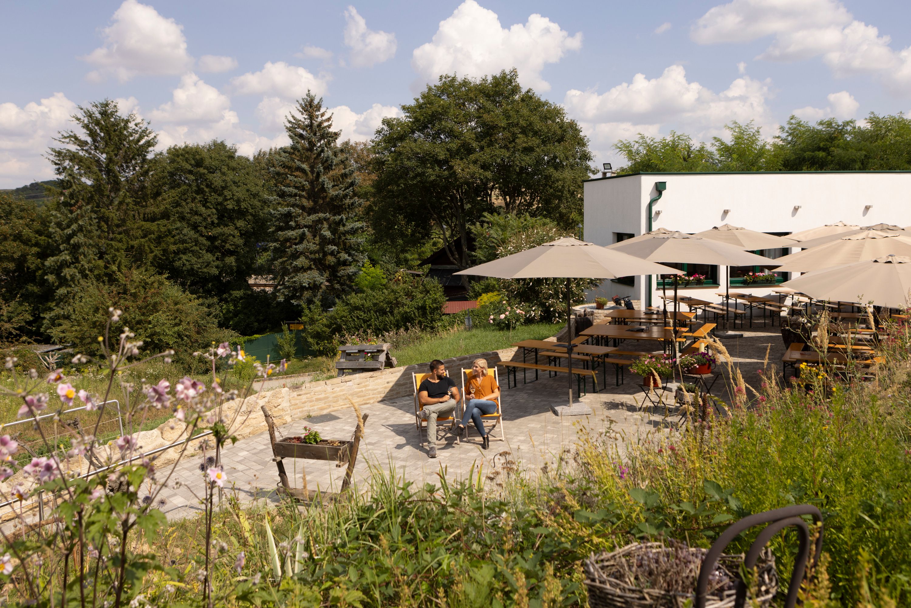 Ein Paar sitzt auf Liegestühlen in einem Garten mit Tischen und Sonnenschirmen, umgeben von Bäumen und Blumen.