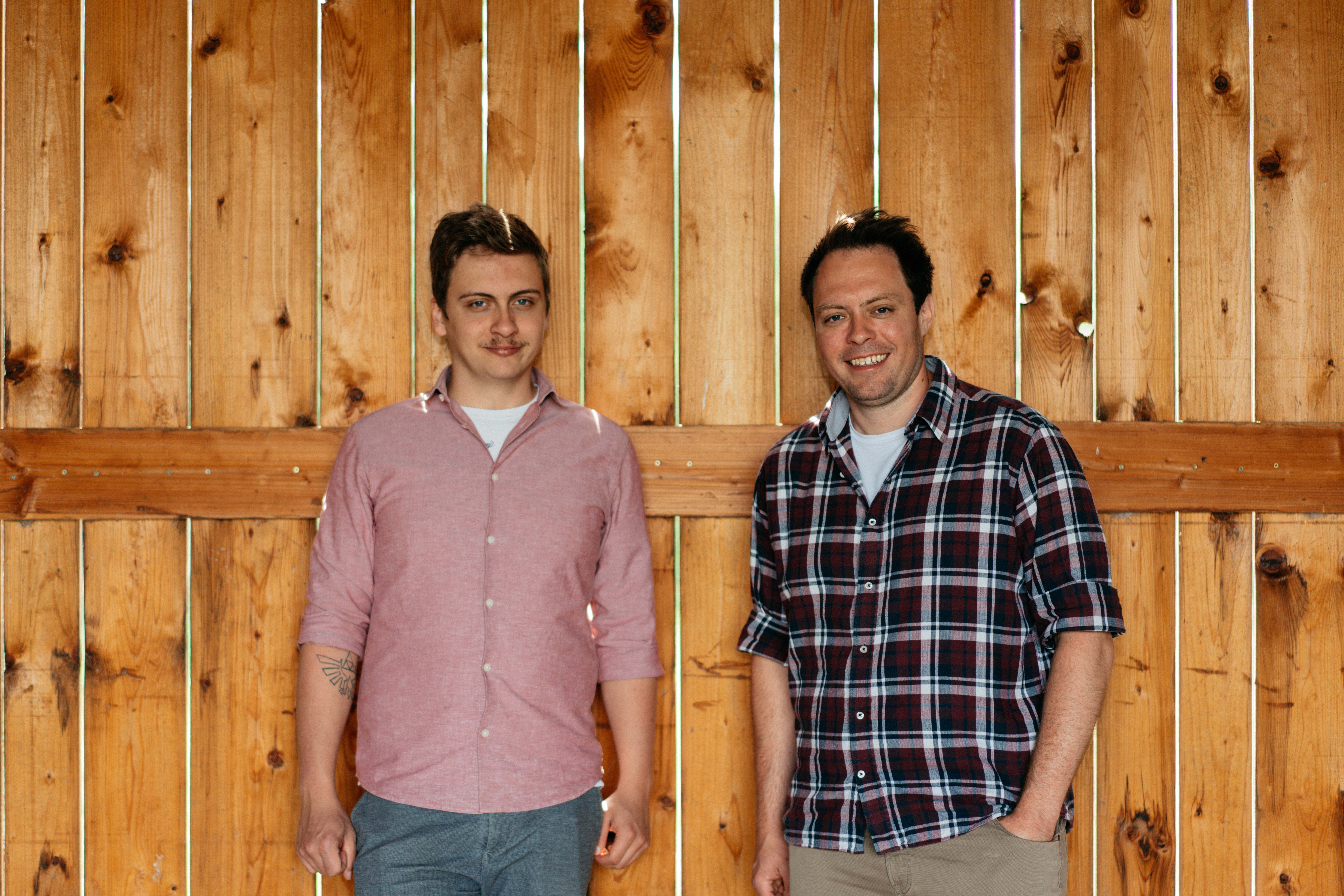Dvaja muži stoja pred drevenou stenou a usmievajú sa do kamery.