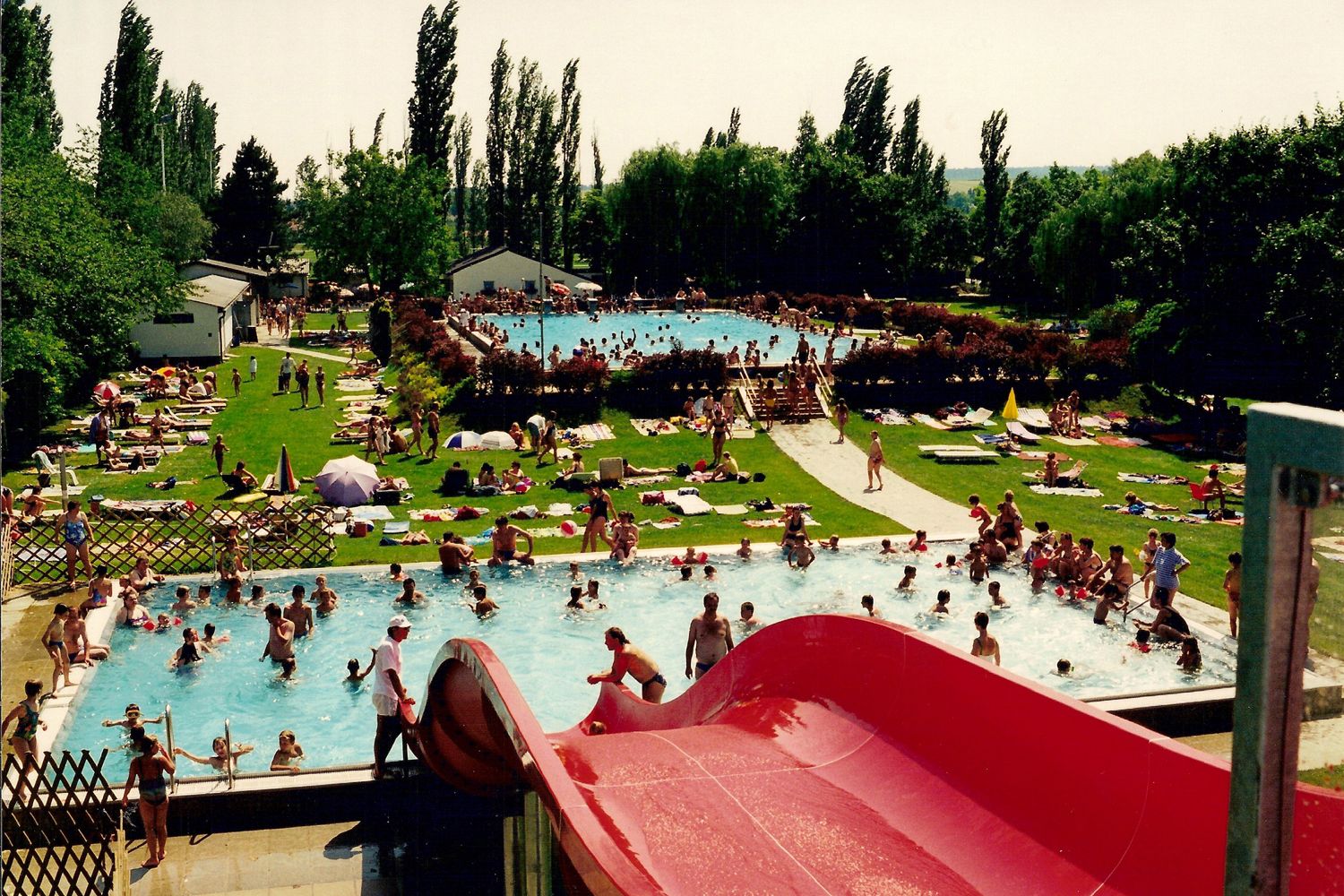 Freibad mit vielen Menschen im Wasser und auf der Liegewiese, rote Rutsche im Vordergrund.