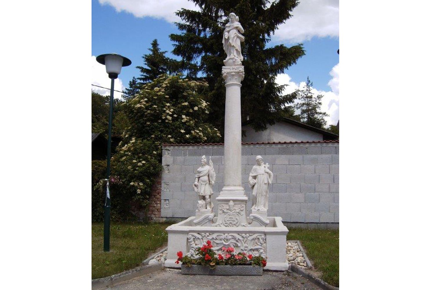 Mariensäule mit Statuen und Blumen, umgeben von Bäumen und einer Mauer.