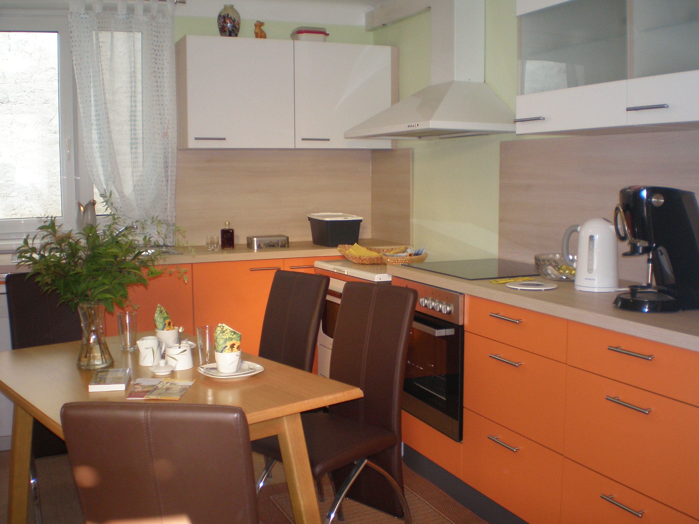 Moderne Küche mit orangefarbenen Schränken, Esstisch mit Stühlen, Kaffeemaschine und Wasserkocher.