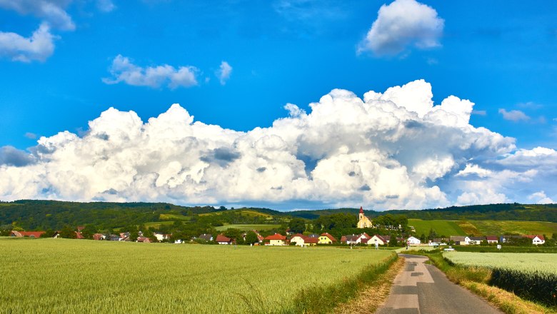 Landschaft mit Dorf, Feldern und Kirche vor großen Wolken.