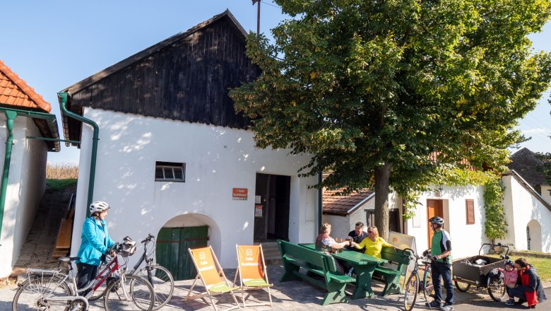 Menschen sitzen auf Bänken vor einem weißen Gebäude mit Fahrrädern in Alberndorf.