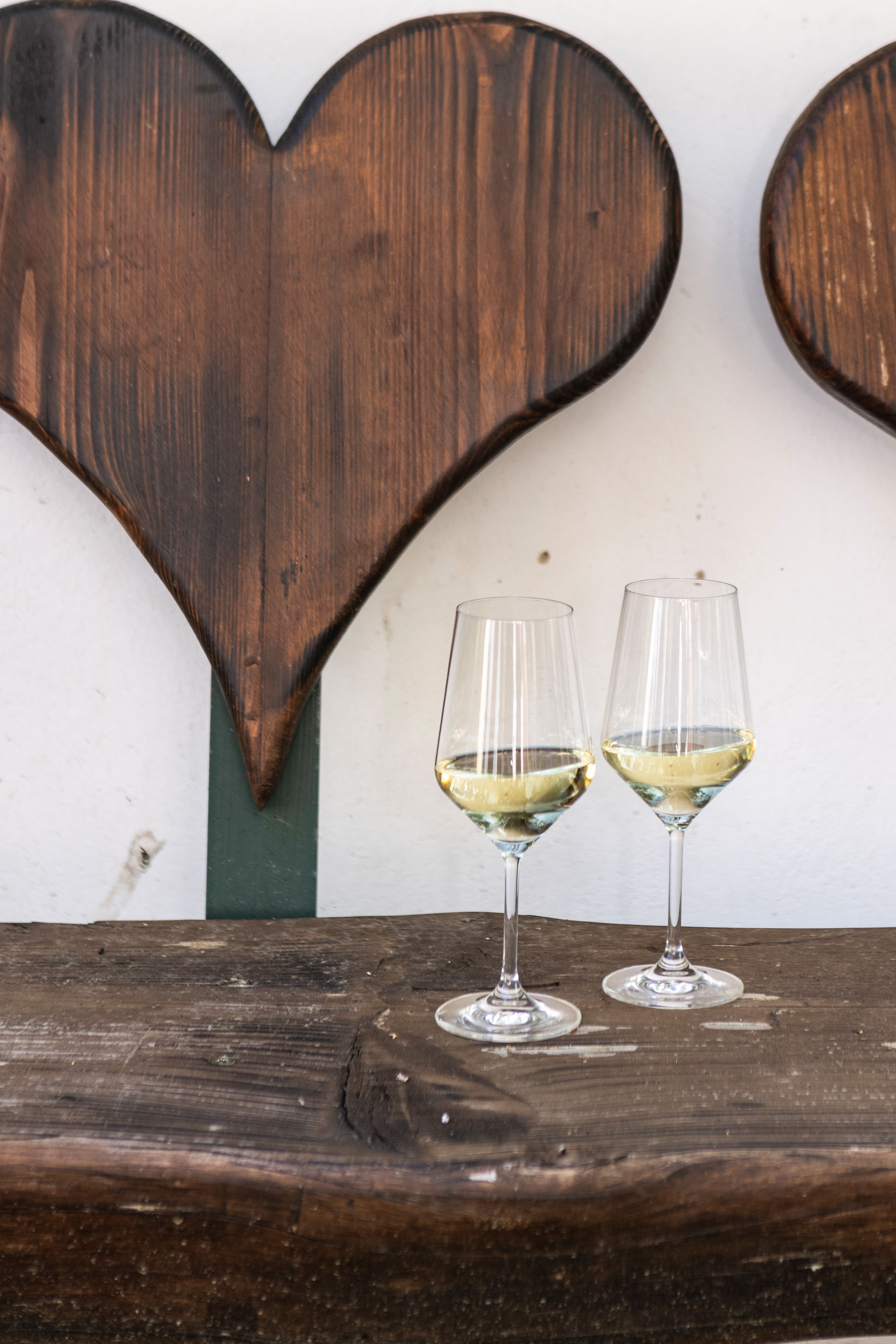 Zwei Weingläser mit Weißwein auf einem Holztisch vor einer Holzwand mit Herzdekor.