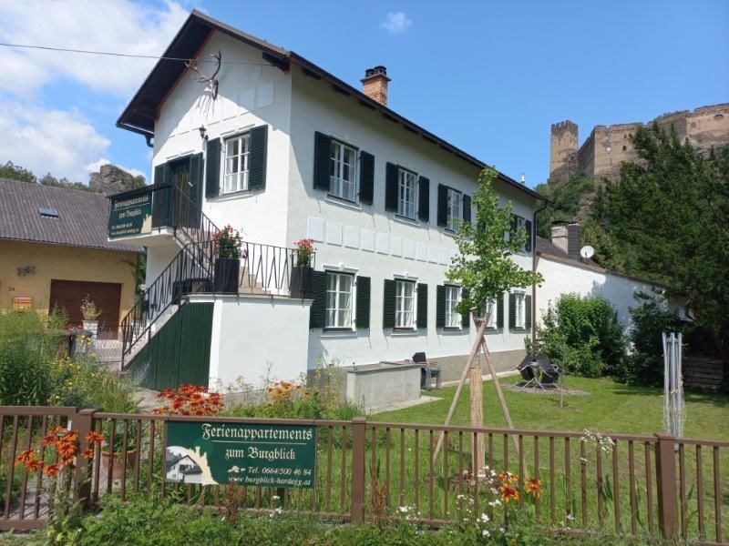 Ein weißes Gebäude mit grünen Fensterläden, umgeben von einem Garten, mit einer Burg im Hintergrund.