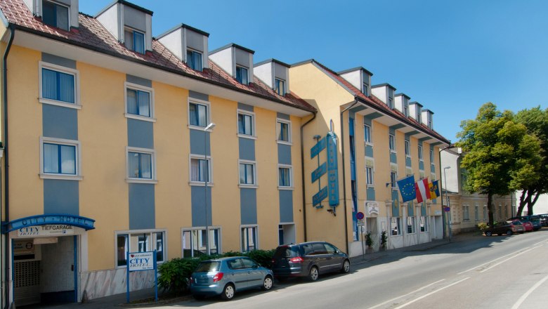 Außenansicht eines gelb-blauen Hotels mit mehreren Fenstern und geparkten Autos davor.