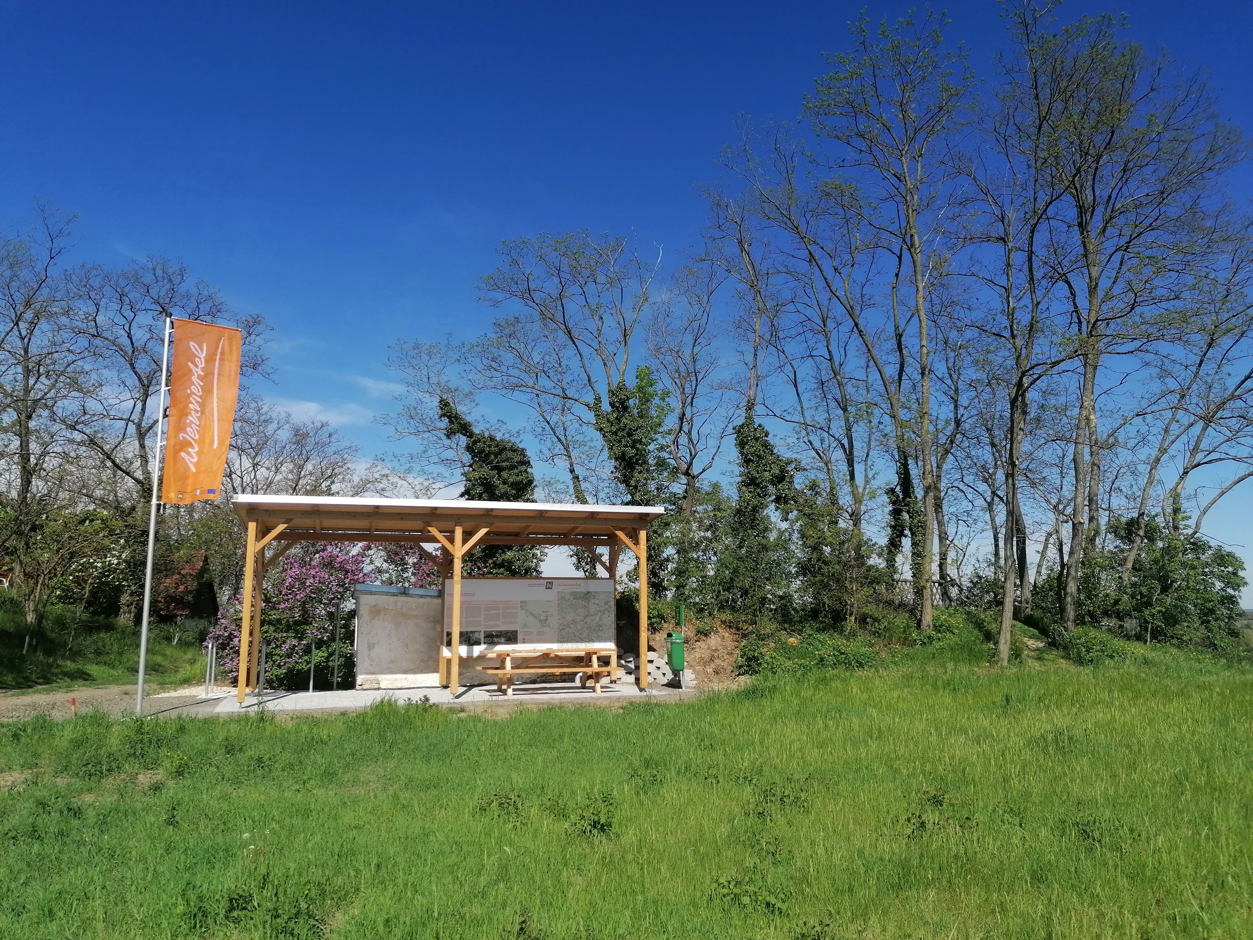 Rastplatz im Weinviertel mit überdachtem Sitzbereich und Fahne, umgeben von Bäumen und Wiese.