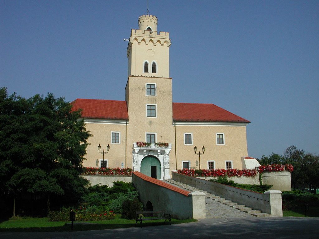 Ein Schloss mit einem Turm und roten Dächern, umgeben von Blumen und Bäumen, unter einem klaren blauen Himmel.