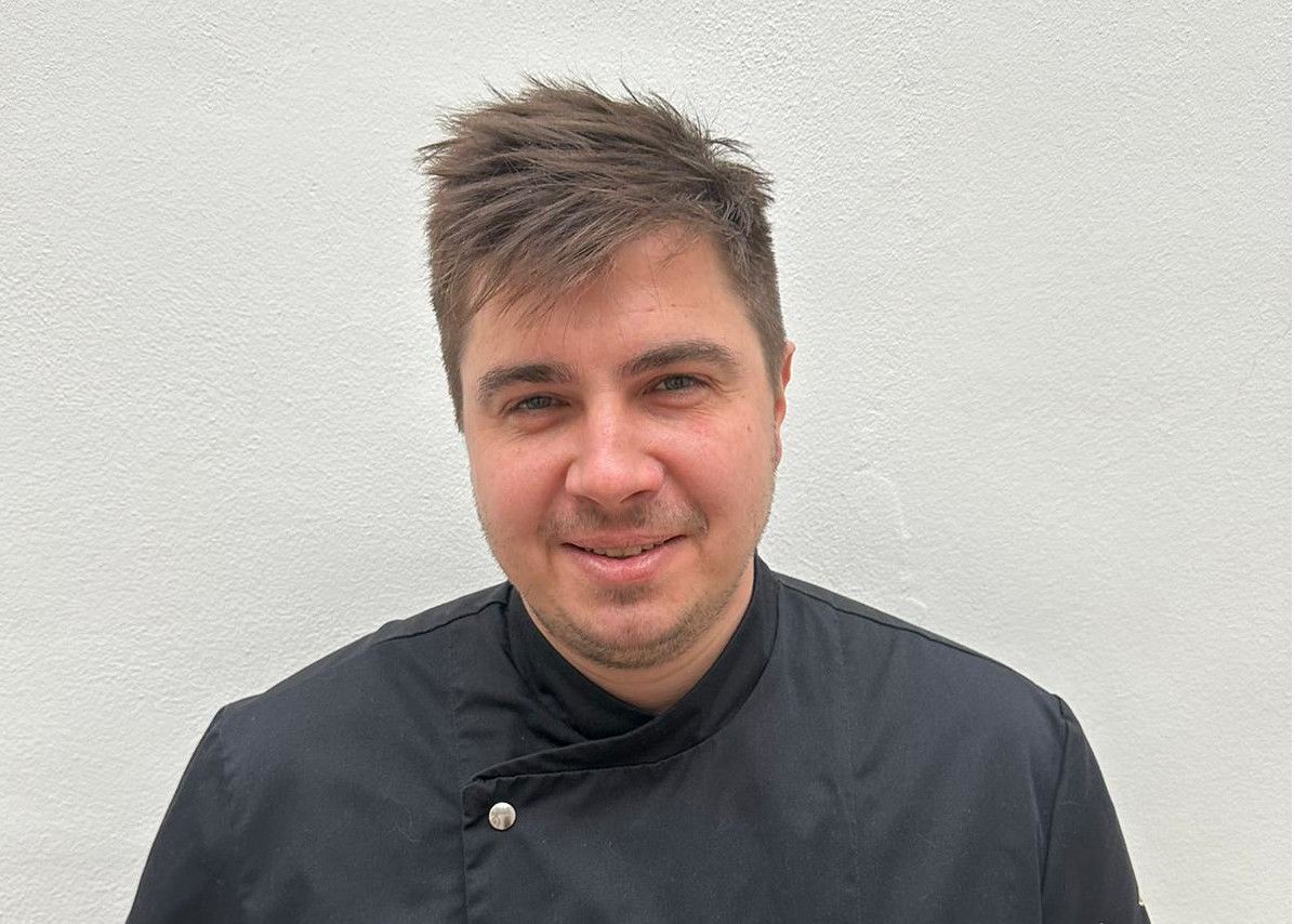 Ein Mann in schwarzer Kochjacke vor einer weißen Wand.