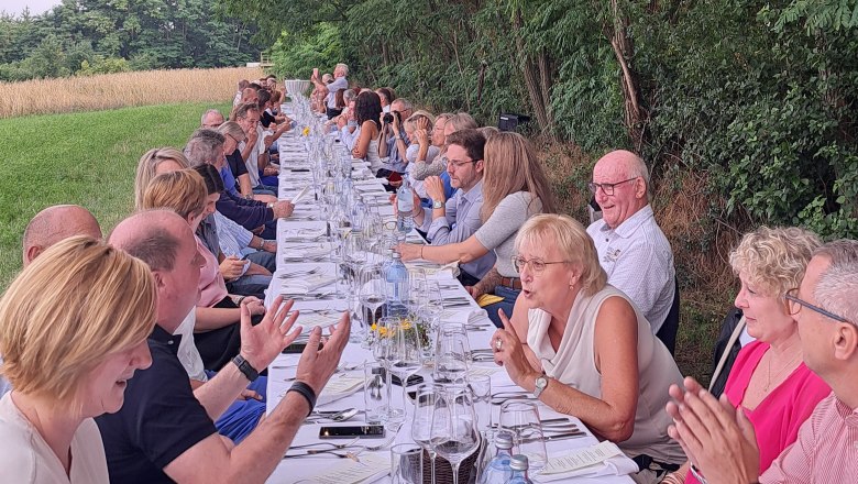 Menschen sitzen an einem langen Tisch im Freien unter Bäumen mit Lichterkette.