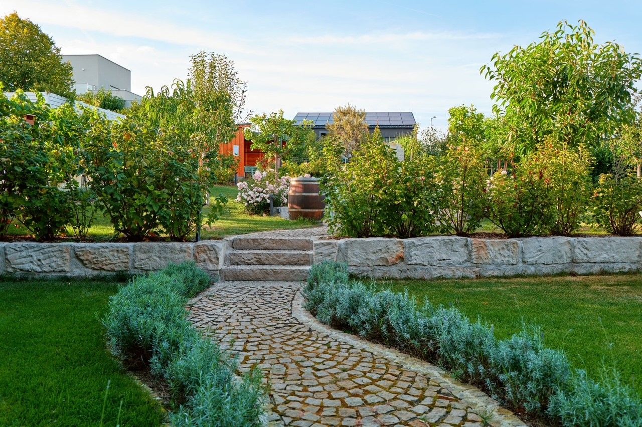 Gepflegter Garten mit Steinweg, Büschen und Bäumen, im Hintergrund ein Gebäude mit Solarpanelen.
