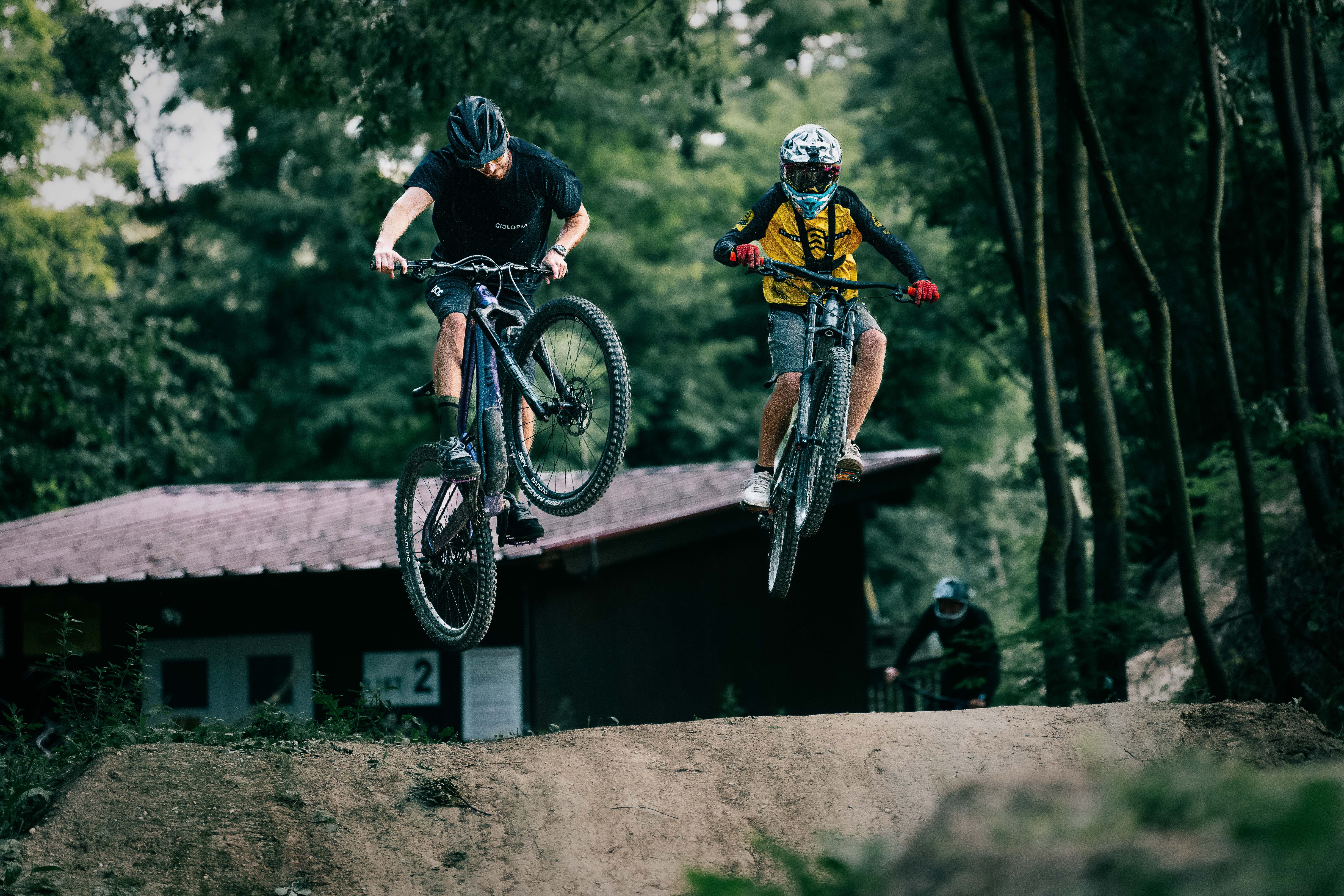 Two mountain bikers jump off a ramp in the woods.