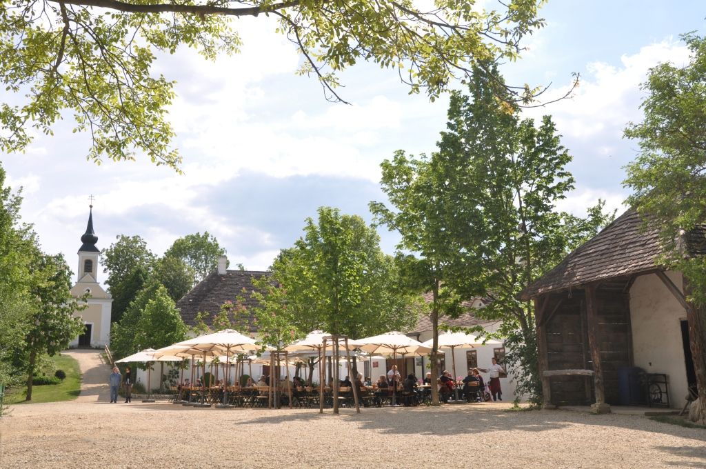 Ein Dorfwirtshaus im Museumsdorf mit Sonnenschirmen und einer Kirche im Hintergrund.