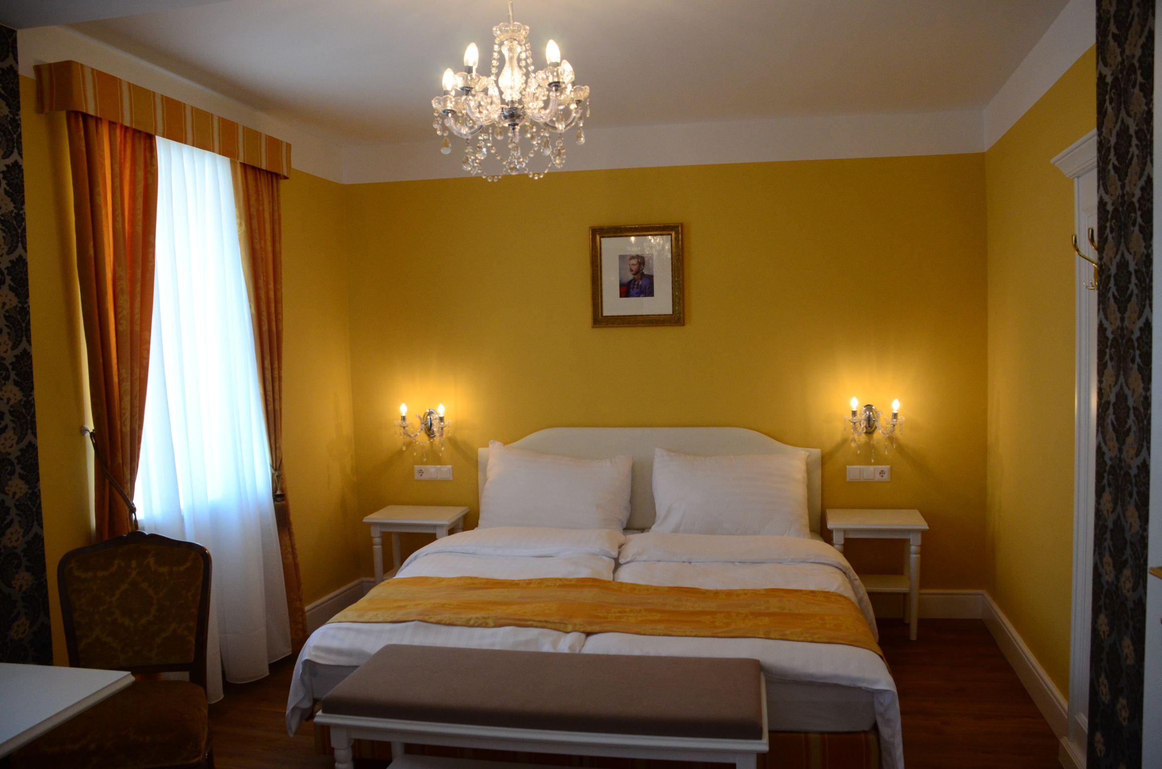 Elegantní hotelový pokoj se žlutými stěnami, manželskou postelí, lustrem a obrazem na stěně.