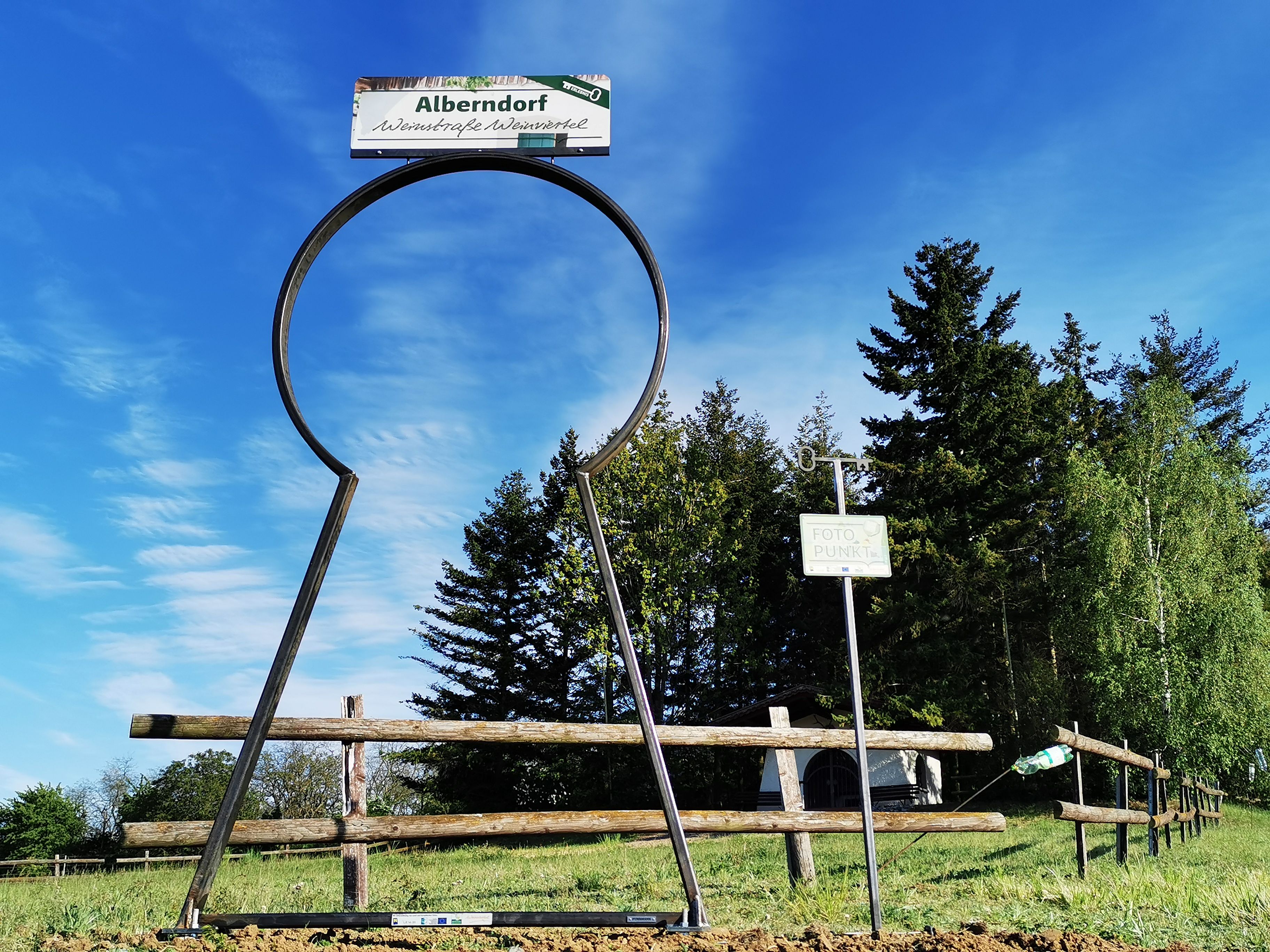 Großer Schlüssellochrahmen mit Schild 'Alberndorf' und 'Foto Punkt' vor Bäumen und blauem Himmel.