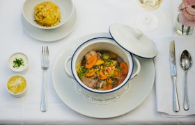 Ein gedeckter Tisch mit einem Eintopf in einem wei&szlig;en Topf, einem Kn&ouml;del, zwei So&szlig;en und einem Glas Wei&szlig;wein.