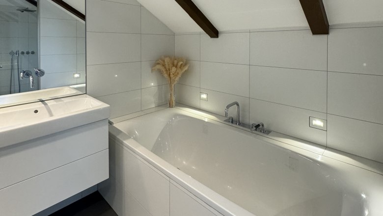 Modernes Badezimmer mit Badewanne, Waschbecken und schr&auml;ger Decke mit Holzbalken.
