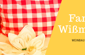 Rotes Kissen mit Karomuster und Handtuch in Blumenform auf einem Bett. Text: Familie Wi&szlig;mann, Weinbau & Privatzimmer.