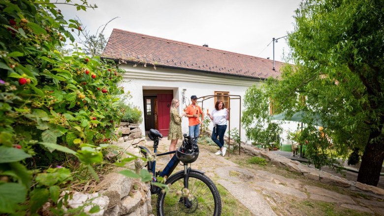 Drei Personen stehen vor einem wei&szlig;en Haus mit rotem Dach, ein Fahrrad im Vordergrund.