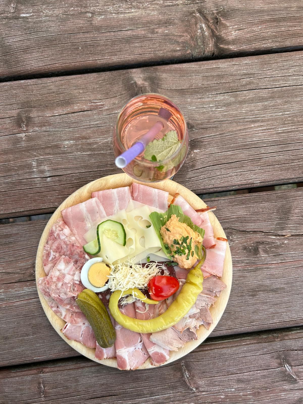 Ein Teller mit Aufschnitt, Käse, Ei, Gurke, Paprika und Brotaufstrich auf einem Holztisch. Daneben ein Glas Roséwein mit Strohhalm.