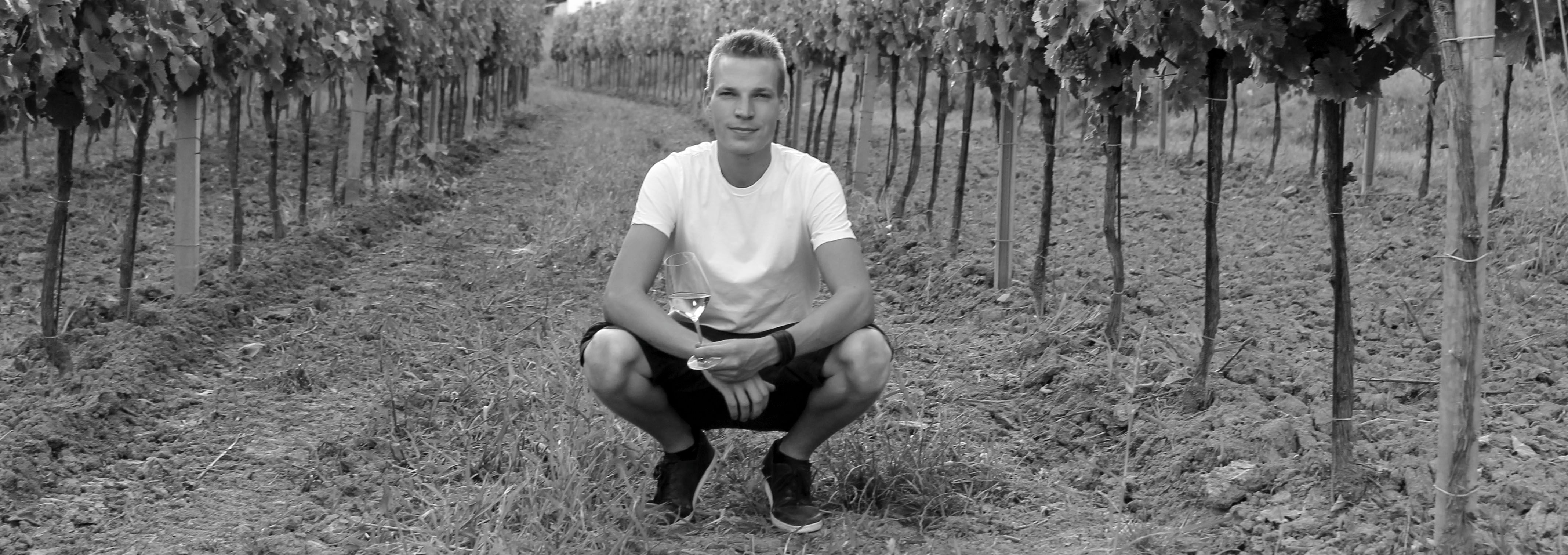 Osoba dřepící na vinici se sklenicí vína v ruce.