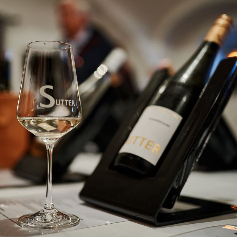 Ein Weinglas mit der Aufschrift 'Sutter' steht vor einer Weinflasche auf einem Tisch.