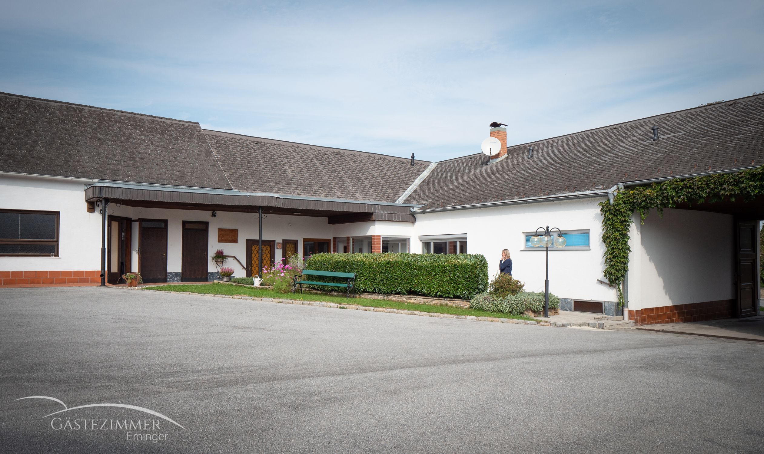 Ein Hof mit einem Gebäude, Parkplätzen und einer Hecke. Eine Person steht neben der Hecke. Auf dem Gebäude ist eine Satellitenschüssel zu sehen.