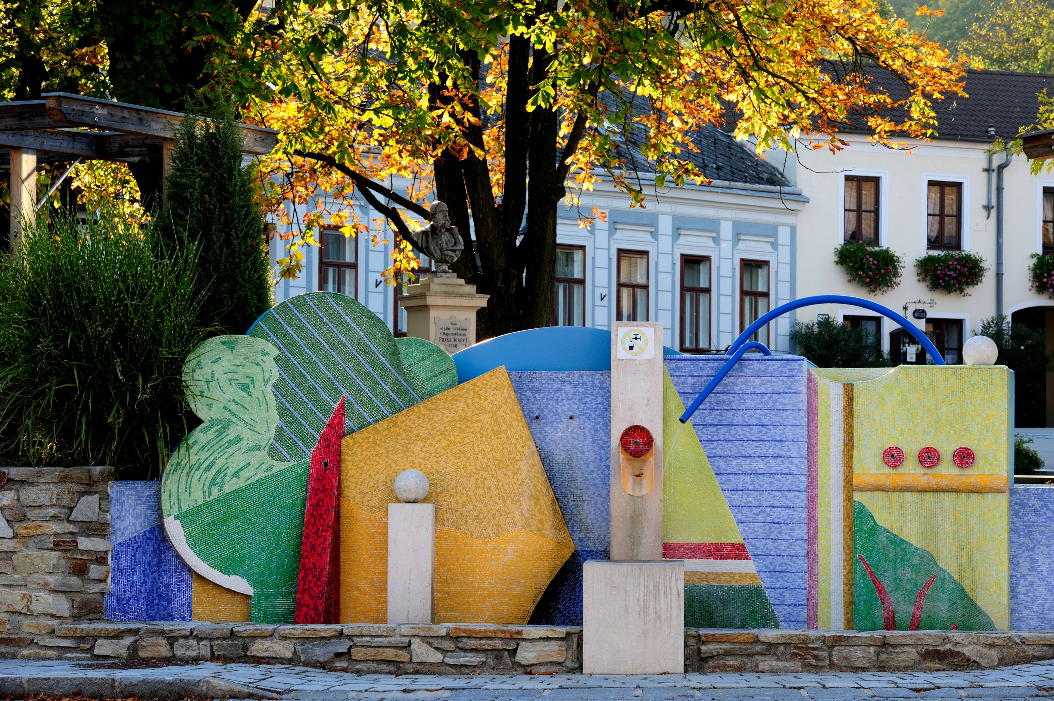 Colorful fountain in Maissau with mosaic decorations and autumn trees in the background.