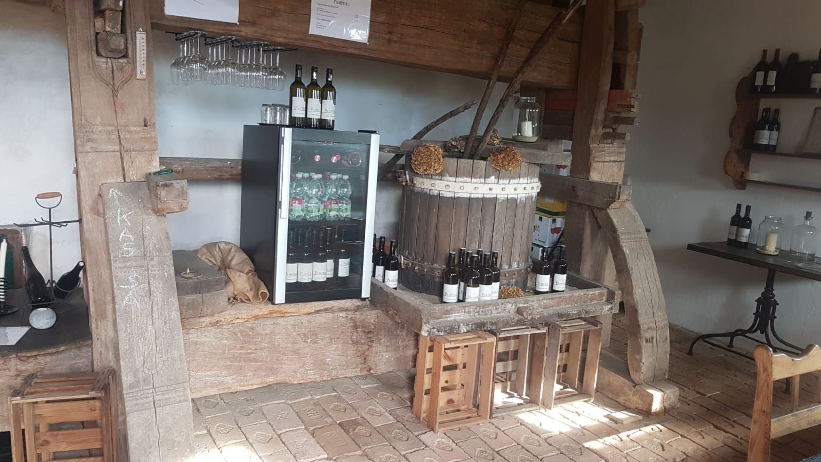 Innenansicht einer rustikalen Weinstube mit Weinflaschen, Gläsern und einem Kühlschrank.