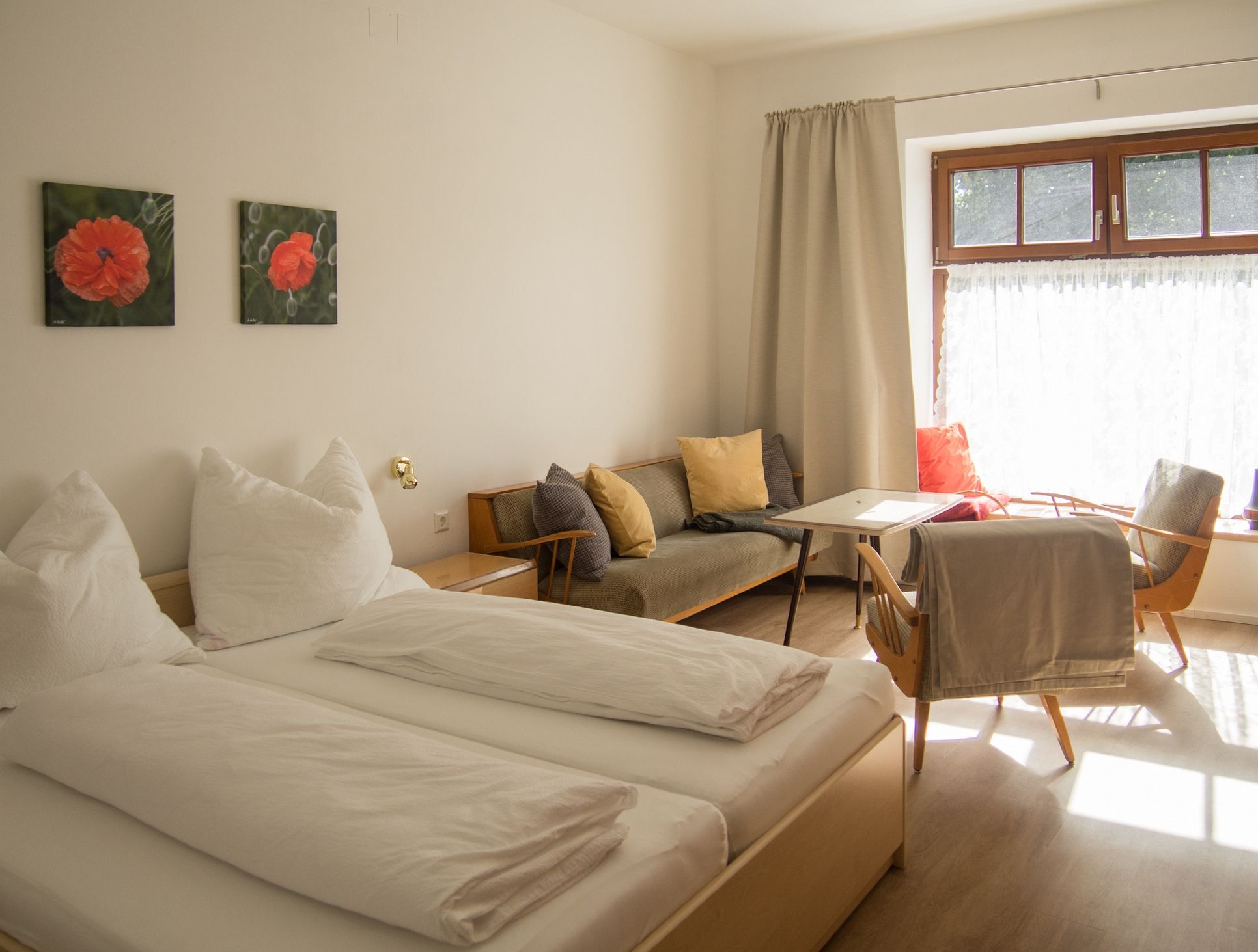 Ein gemütliches Schlafzimmer mit Doppelbett, Sofa, Tisch und Stühlen. Helle Vorhänge und zwei Blumenbilder an der Wand.