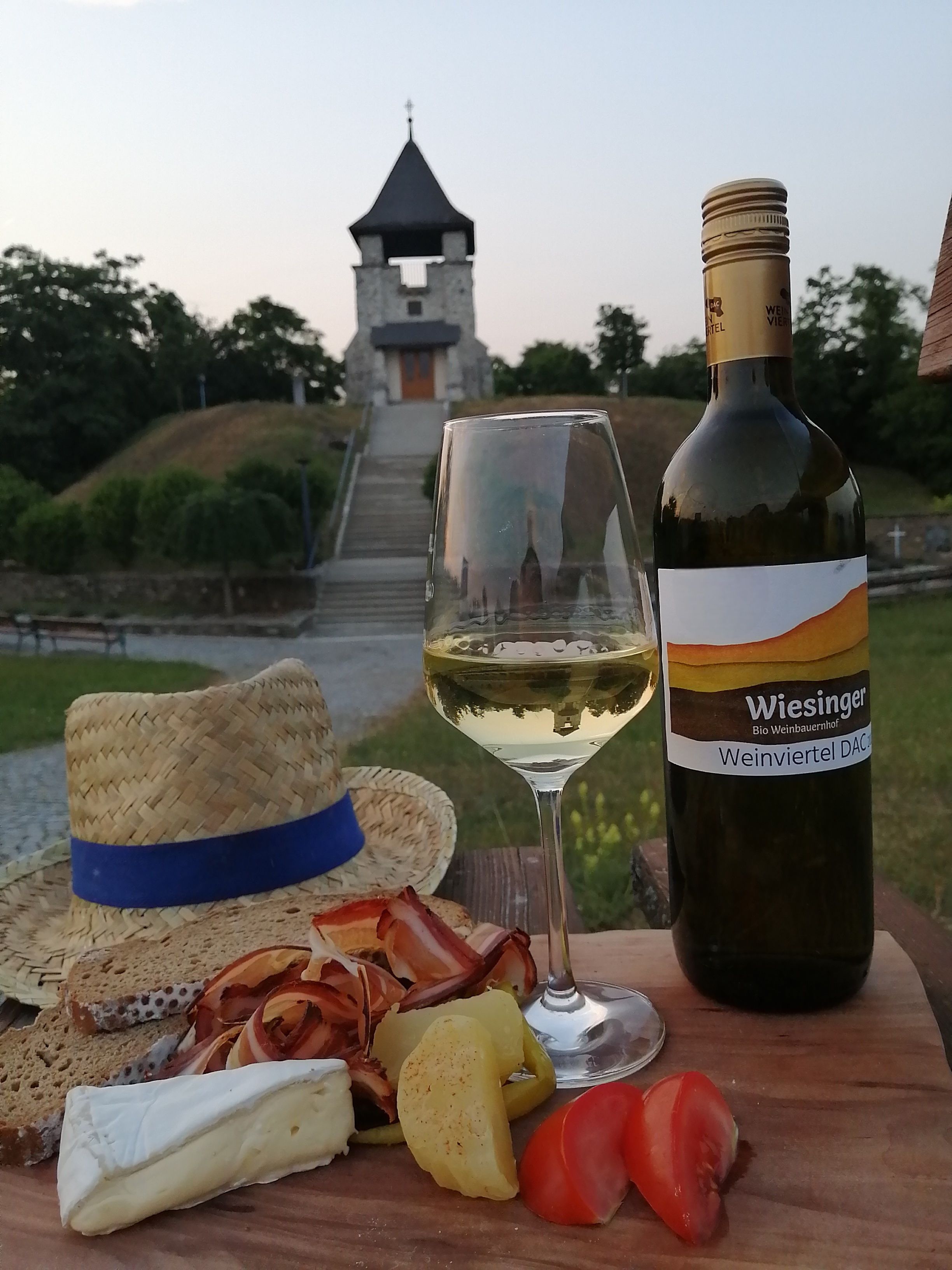 Ein Glas Weißwein und eine Flasche vor einem Turm, mit Käse, Schinken und Brot auf einem Holzbrett.