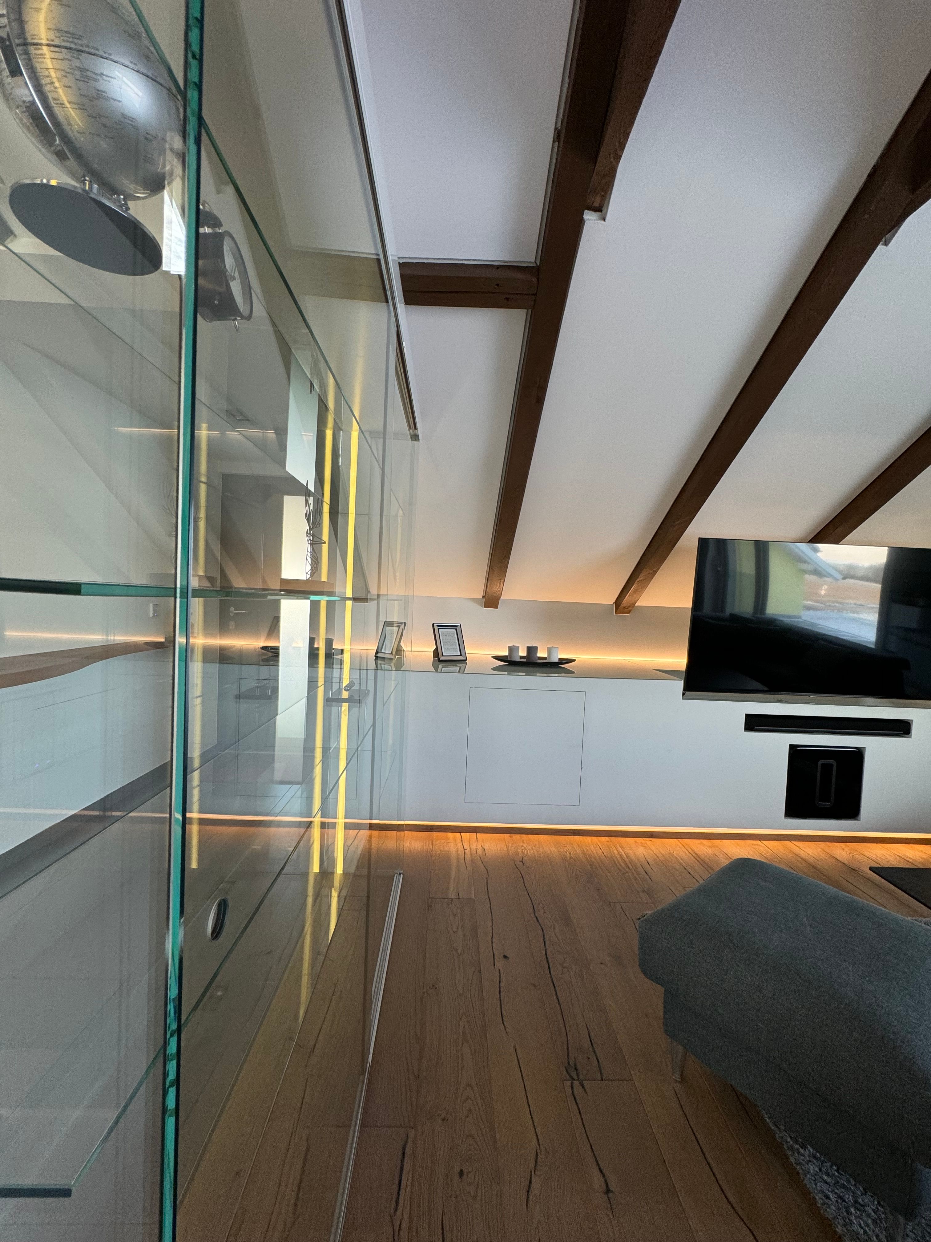 Modernes Wohnzimmer mit Holzboden, Glasvitrine, TV und schräger Decke mit Holzbalken.