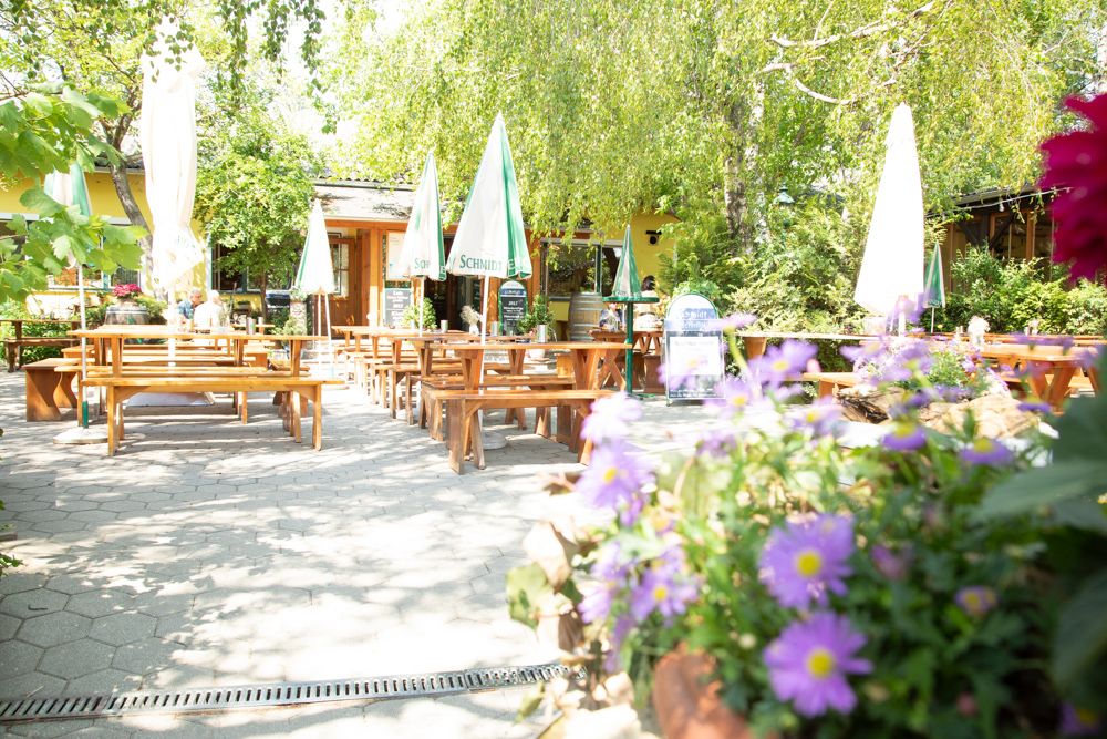 Ein sonniger Gastgarten mit Holzbänken und Tischen, umgeben von grünen Bäumen und bunten Blumen.