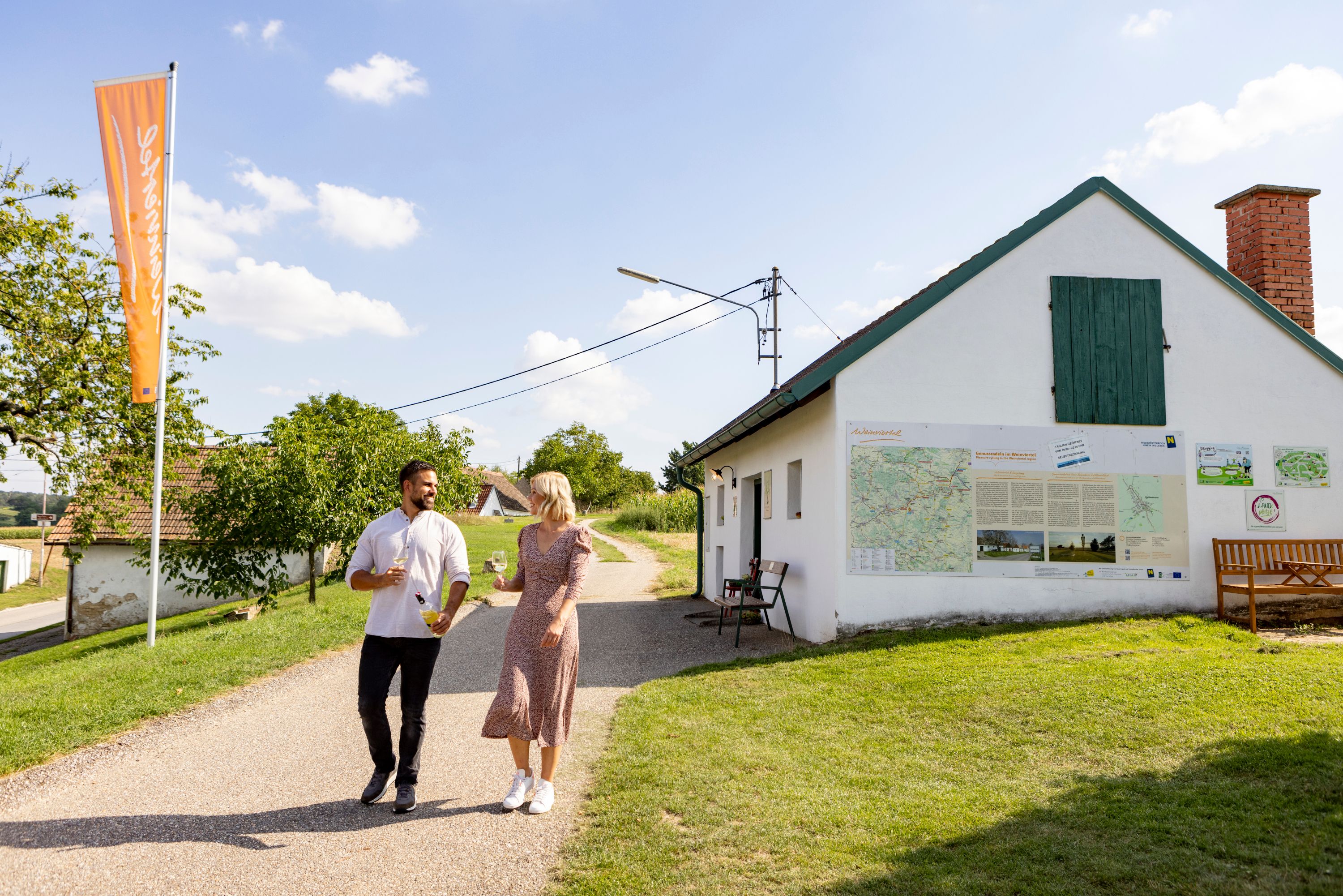 A couple walks past a white building with maps on the wall, wine glasses in hand.