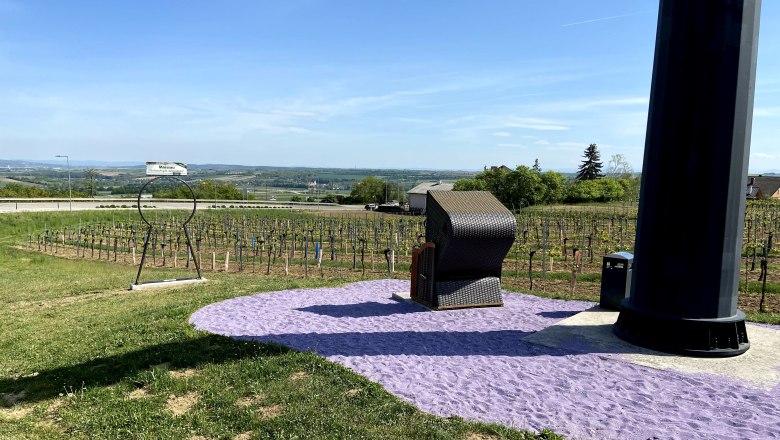 A beach chair stands on purple sand next to a large black pillar, surrounded by vineyards and a vast landscape.