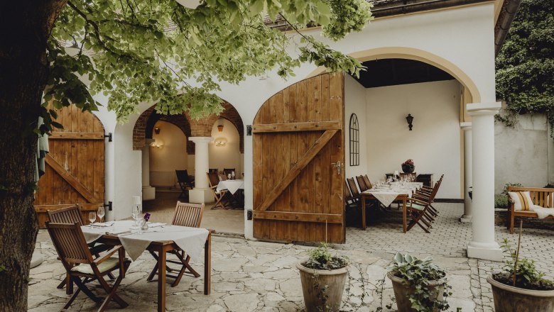 Cozy guest garden with wooden tables and chairs under a tree, surrounded by white walls and open wooden doors.