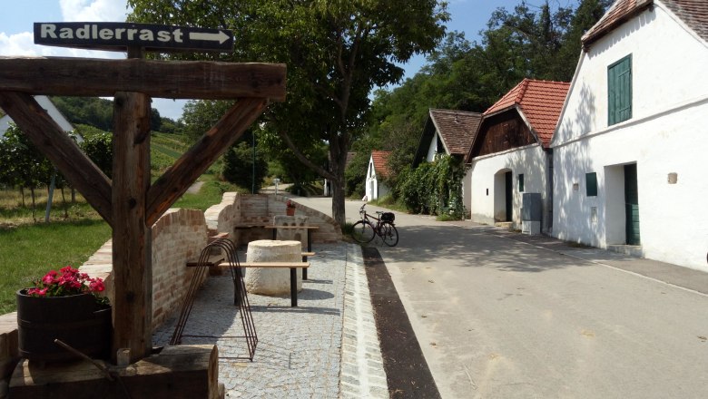 Ein Rastplatz für Radfahrer in Mailberg mit einem Holzschild, das auf die Radlerrast hinweist. Im Hintergrund sind Weinkeller und ein Fahrrad zu sehen.