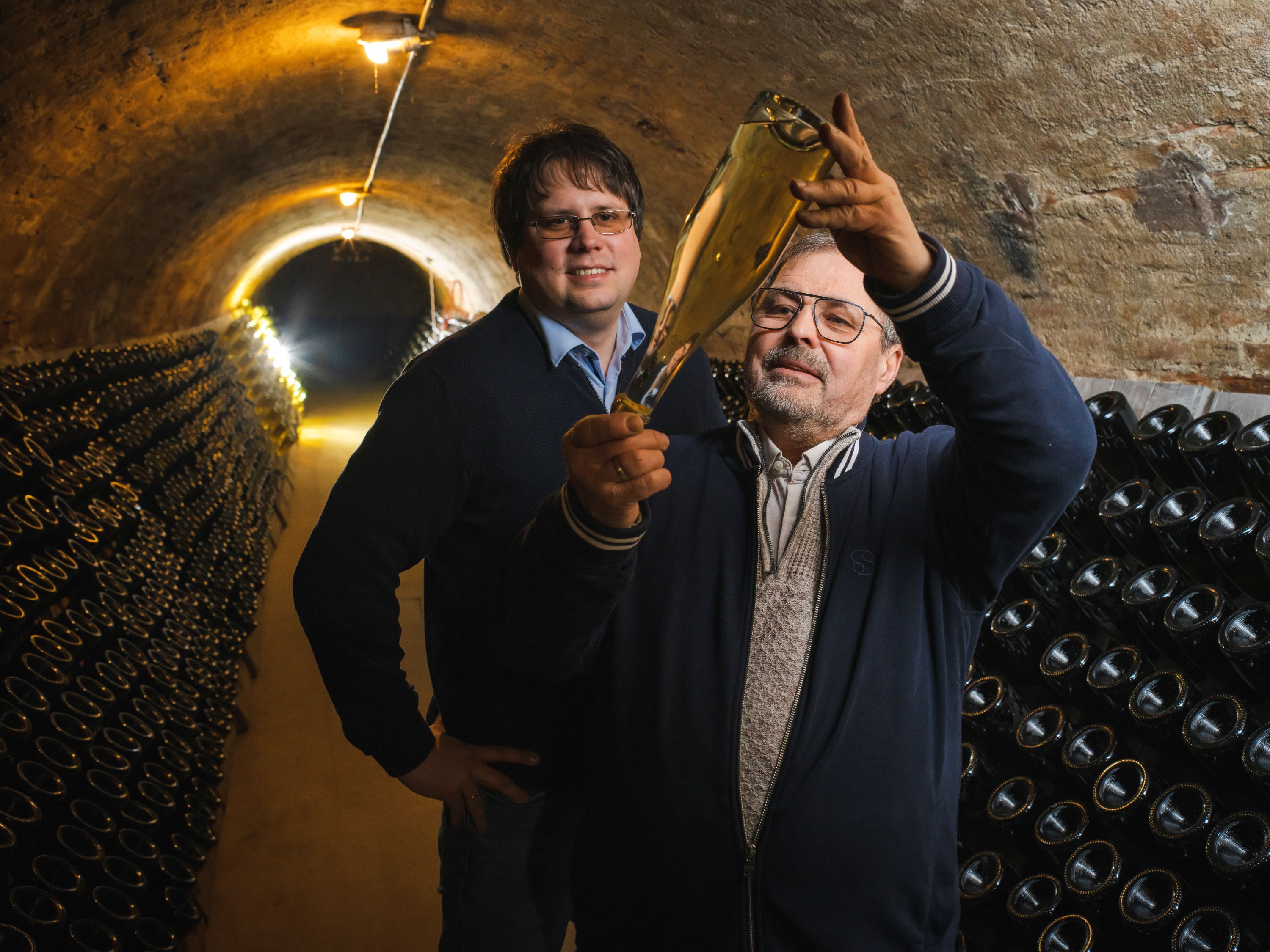 Zwei männliche Sektproduzenten sind bei der Kontrolle des Rüttelvorgangs in einem Keller mit Flaschen zu sehen.