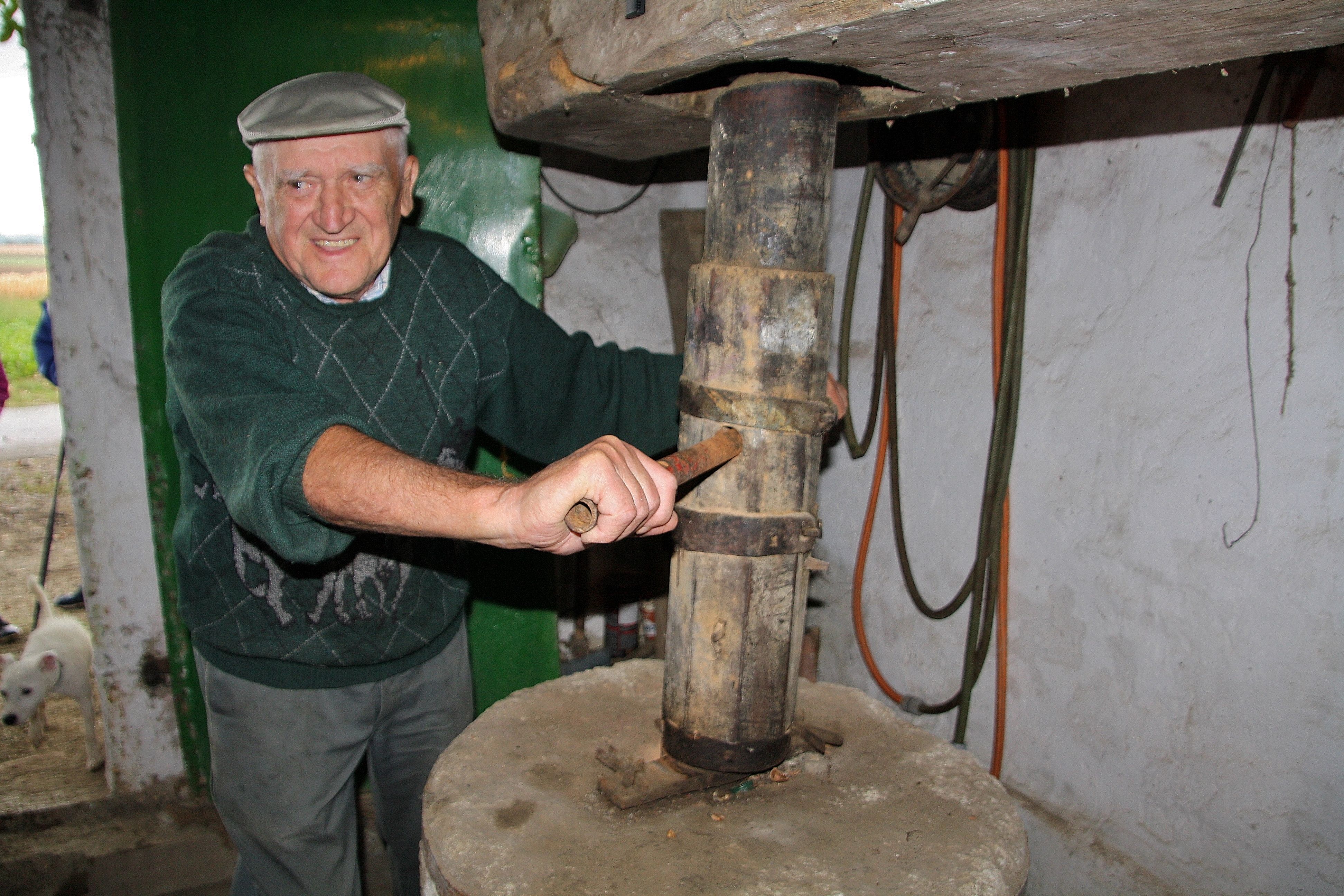 Ein älterer Mann mit Mütze bedient eine alte mechanische Presse in einem rustikalen Raum.