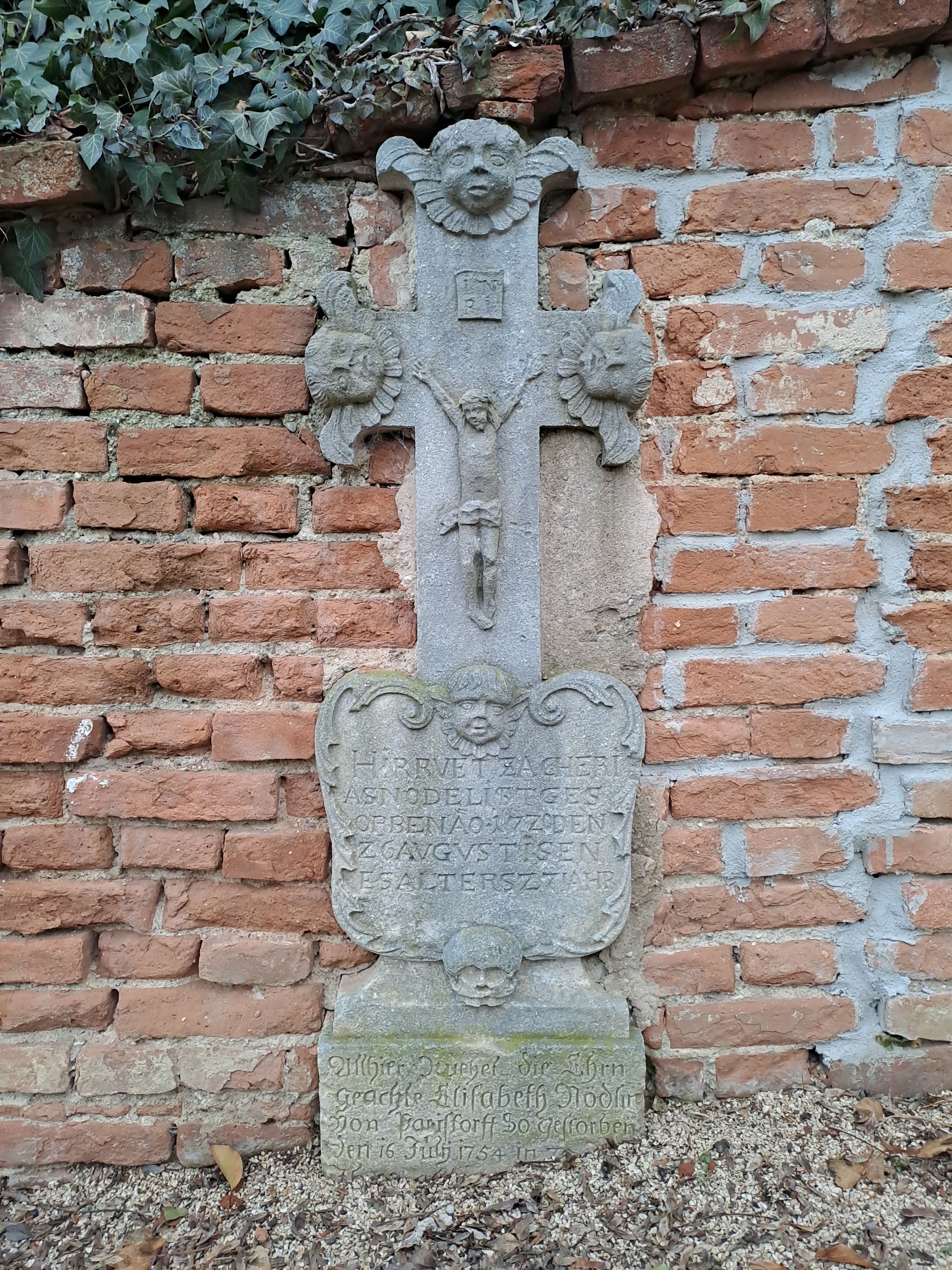 A sandstone cross with inscription and decorations in front of a brick wall.