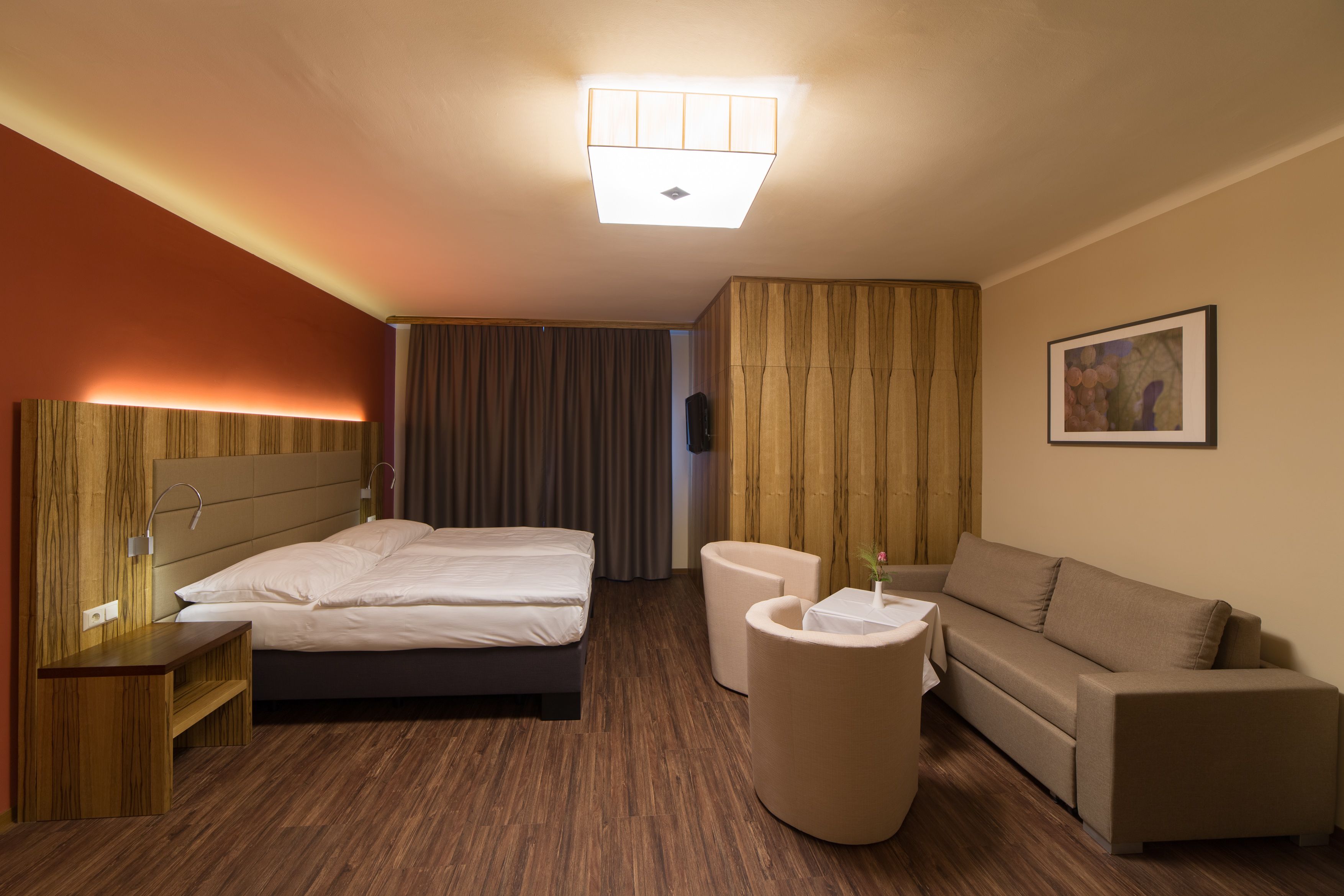 Modern eingerichtetes Hotelzimmer mit Doppelbett, Sofa, zwei Sesseln und einem kleinen Tisch. Holzboden und warme Beleuchtung.