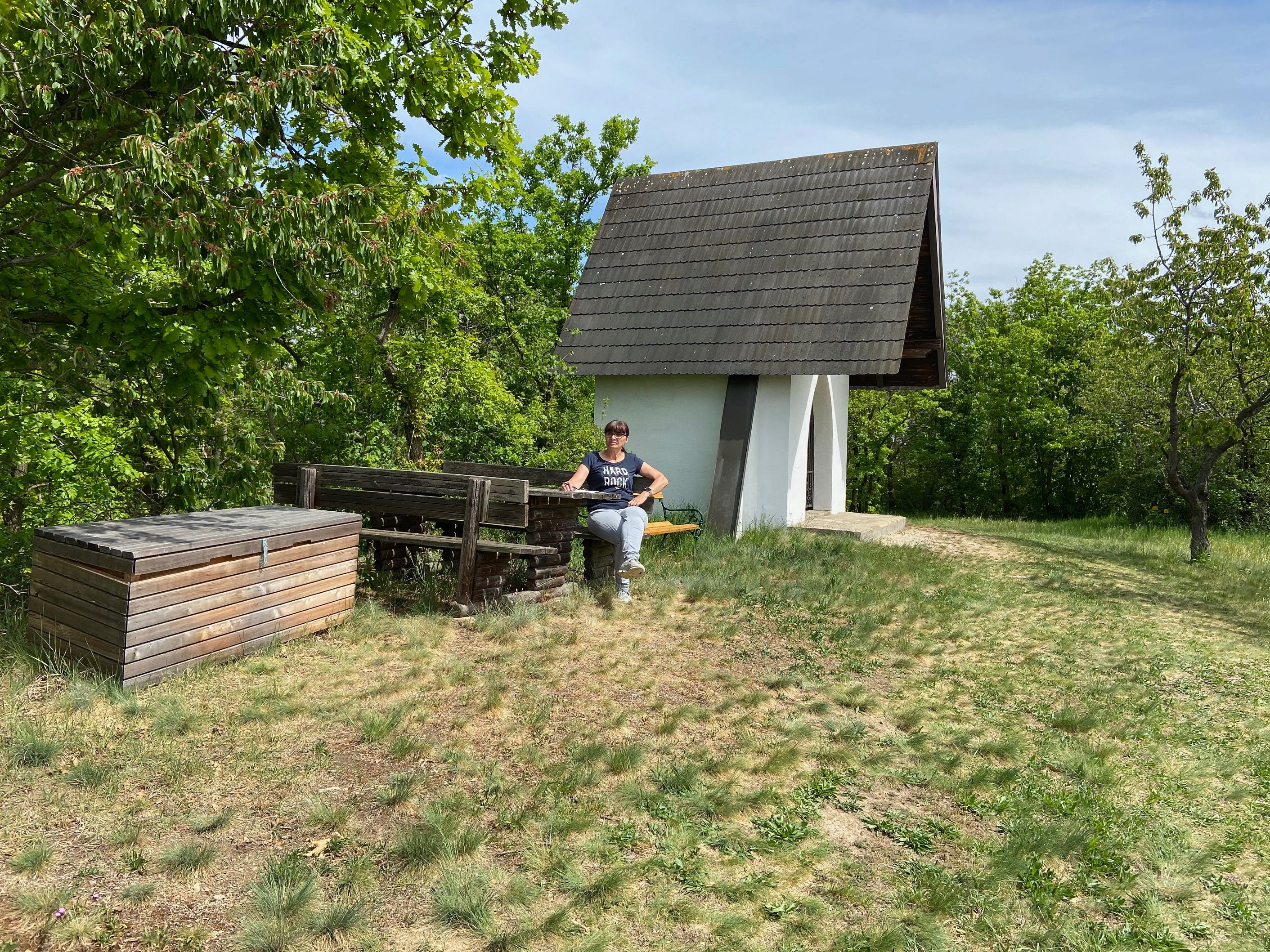 Eine kleine weiße Kapelle mit spitzem Dach in einer grünen Landschaft, daneben eine Person auf einer Bank.