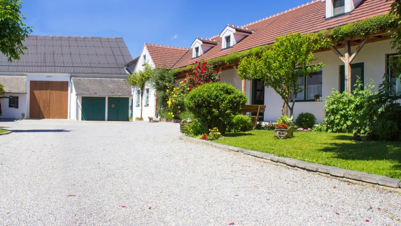 Urlaubsweingut, © Fam. Lobner Ein idyllisches Weingut mit Kiesweg, blühenden Gärten und traditionellen Gebäuden unter blauem Himmel.