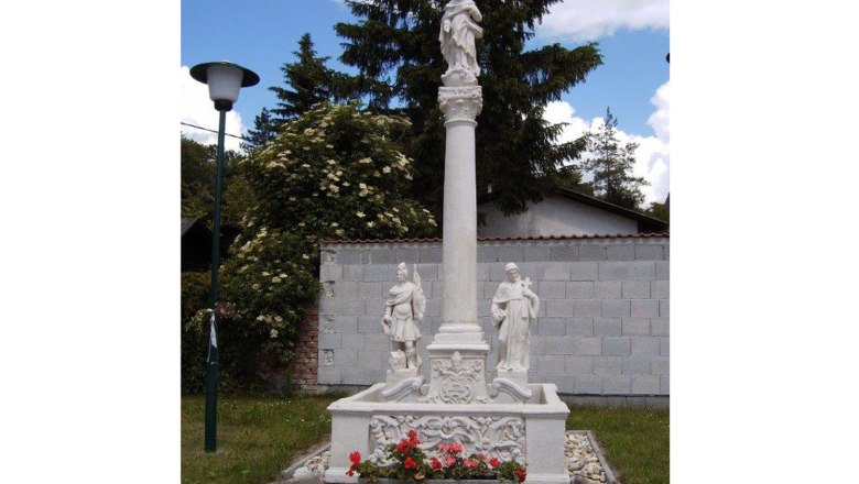 Marian column with statues and flowers, surrounded by trees and a wall.