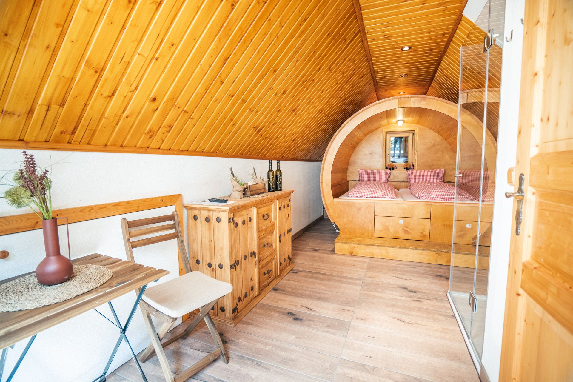Gemütlicher Holzinnenraum im Winzerhof Küssler mit schräger Holzdecke, kleinem Tisch, Schrank und einem runden Bett mit rot-weiß karierten Kissen.
