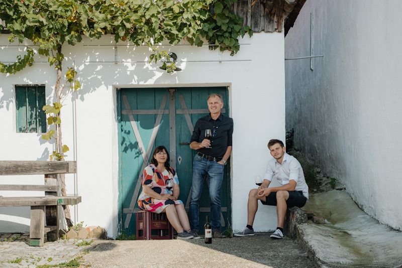 Three people in front of a wine cellar with a blue door and grapevines.